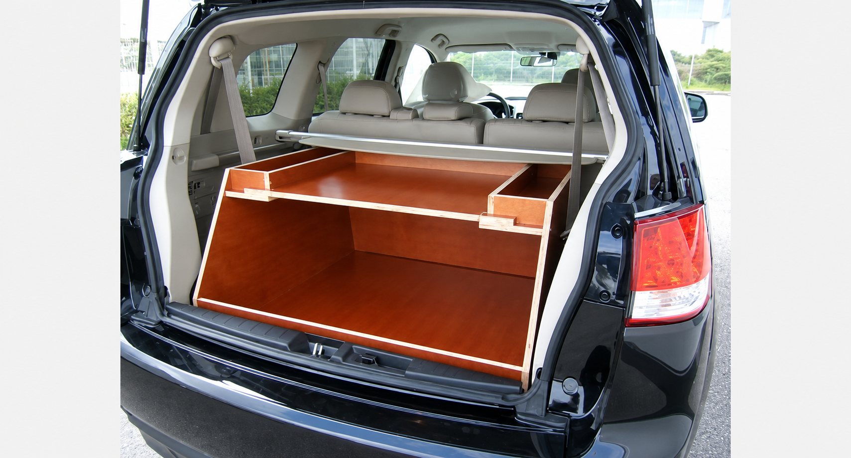 Un meuble de rangement en bois verni à deux niveaux est installé dans le coffre ouvert d'un SUV noir aux sièges beiges.