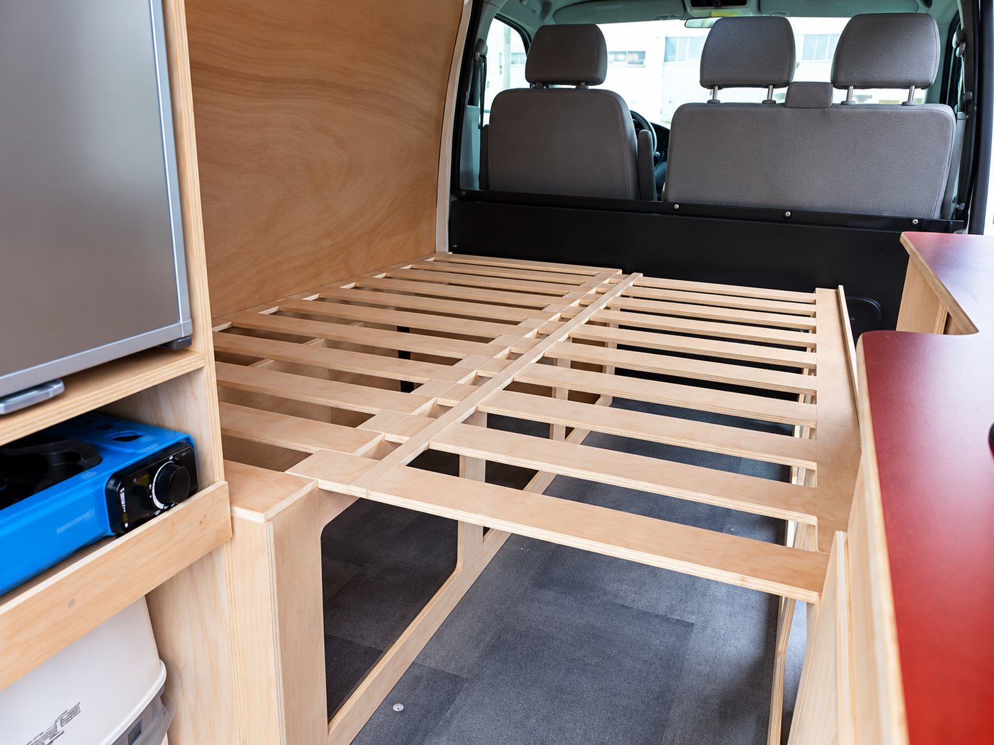 Intérieur d'un van aménagé montrant une structure de lit à lattes en bois clair, un réchaud bleu et un meuble sur le côté, avec les sièges avant visibles au fond.