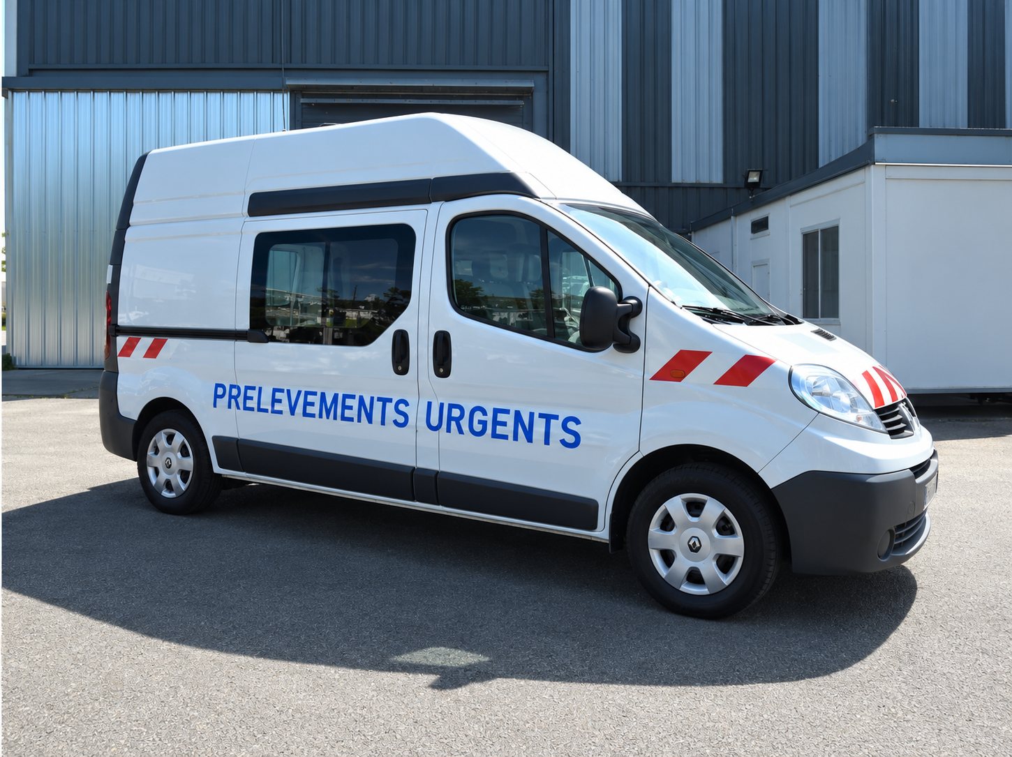 Une camionnette blanche portant l'inscription « Prélèvements urgents » est garée devant un bâtiment industriel.