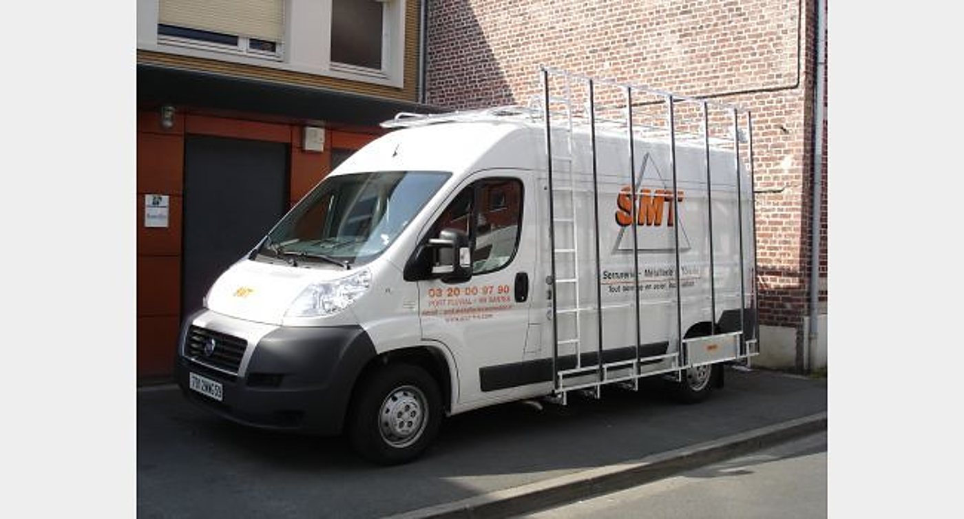 Une camionnette blanche floquée SMT, équipée d'un galerie et d'échelles latérales, est garée devant un mur de briques rouges.