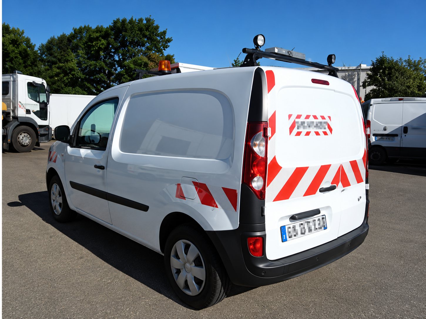 Une camionnette utilitaire blanche équipée de bandes de signalisation rouges et blanches à l'arrière, garée sur un parking à côté d'autres véhicules et d'arbres.