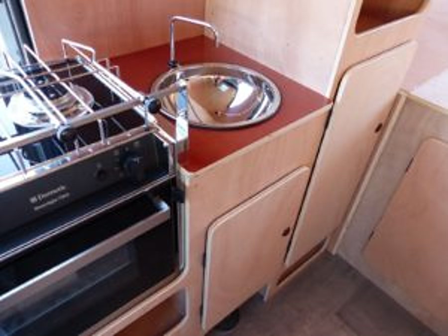 Coin cuisine d'un véhicule aménagé avec plan de travail rouge, évier rond en inox, réchaud à gaz et placards en bois clair.