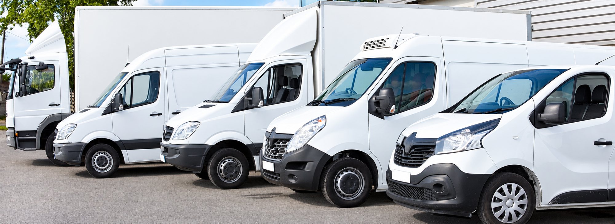 Cinq véhicules utilitaires blancs, camionnettes et camions, sont alignés côte à côte sur un parking devant un bâtiment.