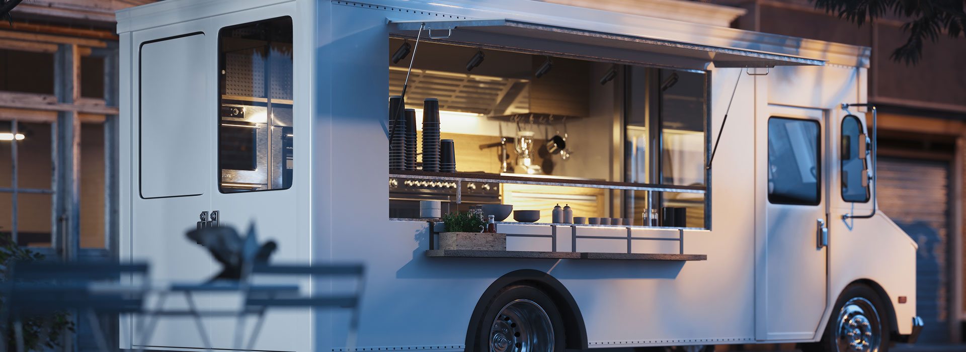 Un food truck blanc avec son volet relevé laisse voir une cuisine éclairée et des gobelets noirs empilés, garé dans la rue au crépuscule.