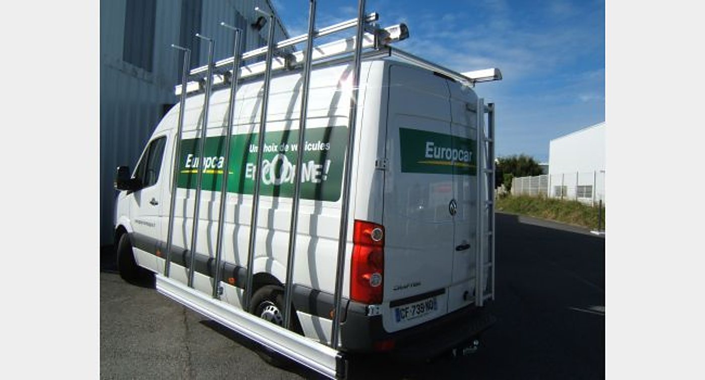 Un fourgon blanc Europcar garé sur un parking, équipé d'une galerie de toit en aluminium et d'une échelle arrière.
