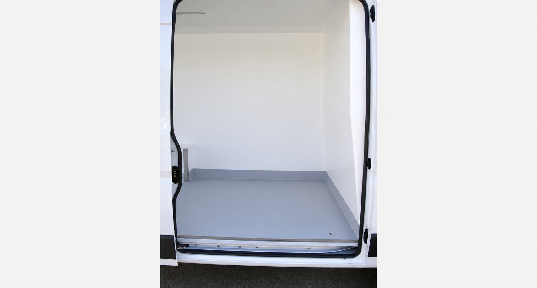 Vue de l'intérieur d'un fourgon utilitaire dont la porte latérale est ouverte, montrant des parois blanches et un plancher gris recouvert d'un revêtement protecteur.