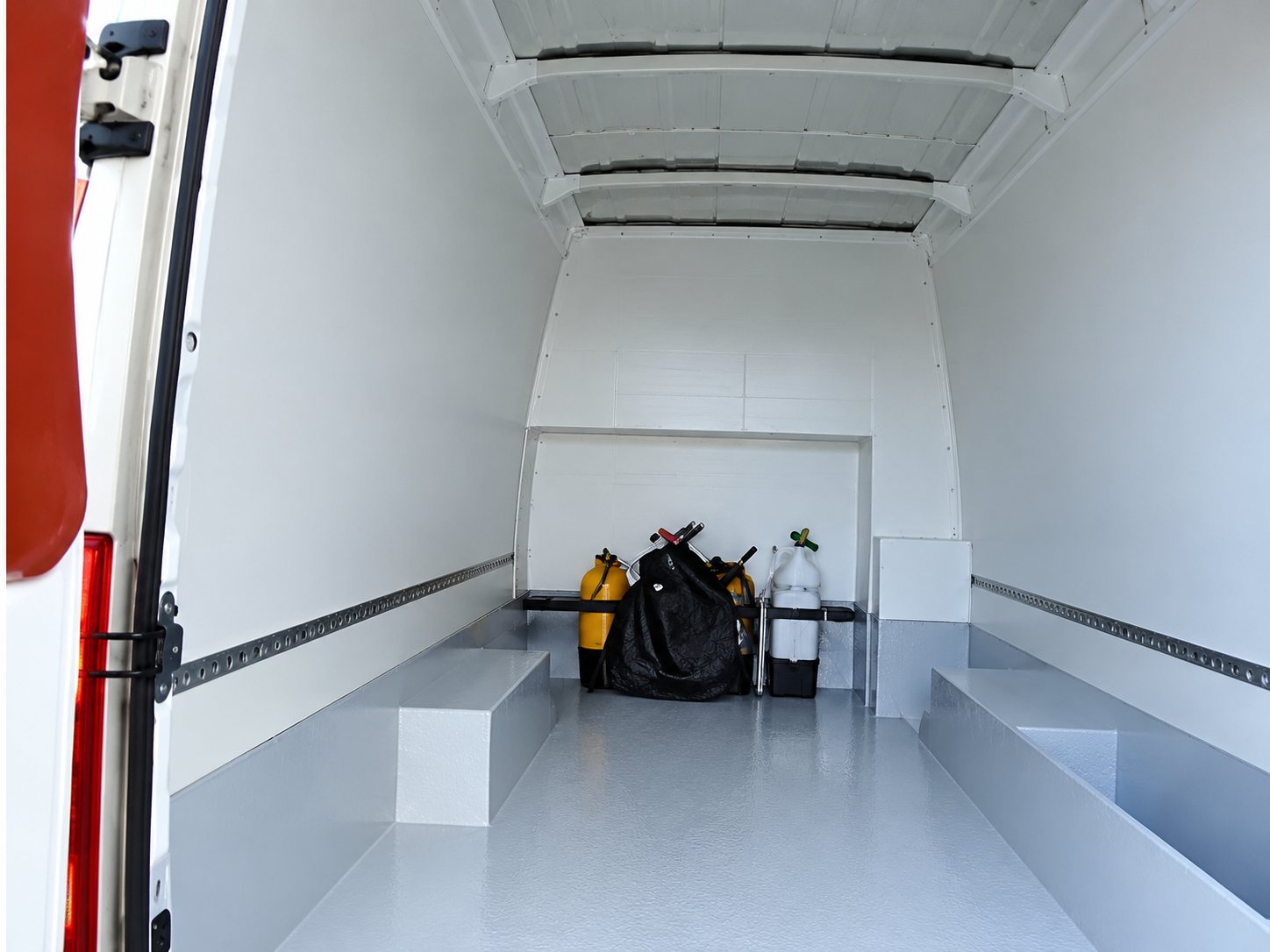 Vue de l'intérieur d'un fourgon utilitaire blanc, vide, avec quelques équipements noirs et des bouteilles placés au fond de l'espace de chargement.