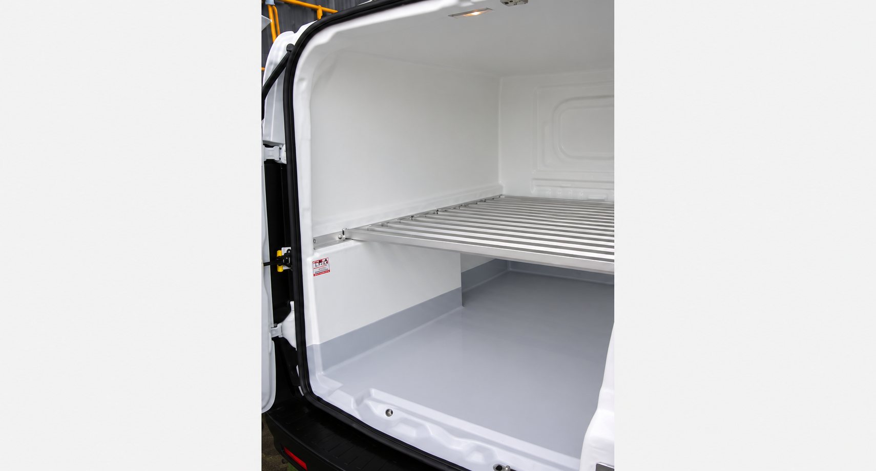 Vue de l'intérieur d'une camionnette blanche aux parois isolées, avec un sol gris et une étagère en aluminium fixée sur le côté.