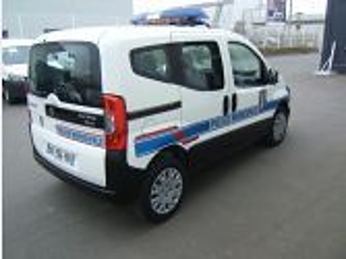 Un petit véhicule utilitaire blanc à bandes bleues et rouges est garé sur un parking devant un bâtiment, vu de l'arrière gauche.