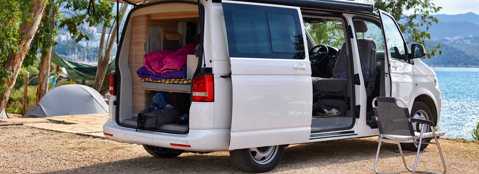 Un van aménagé blanc aux portes ouvertes, avec couchage visible à l'arrière, stationné près d'une tente et d'une chaise pliante au bord d'un lac entouré de montagnes.
